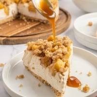 slice of apple crisp cheesecake on a white plate with caramel being poured over the top.