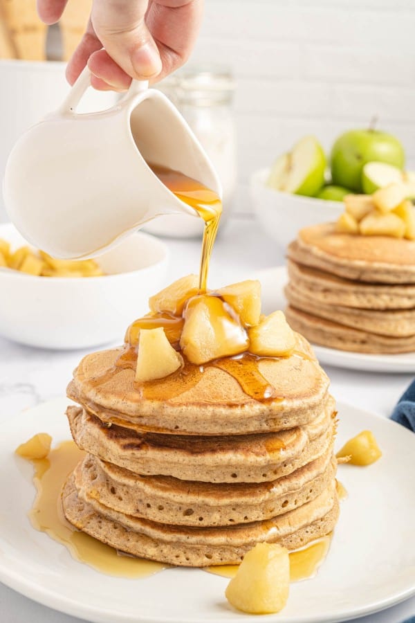 Pouring syrup onto a stack of pancakes with apples.