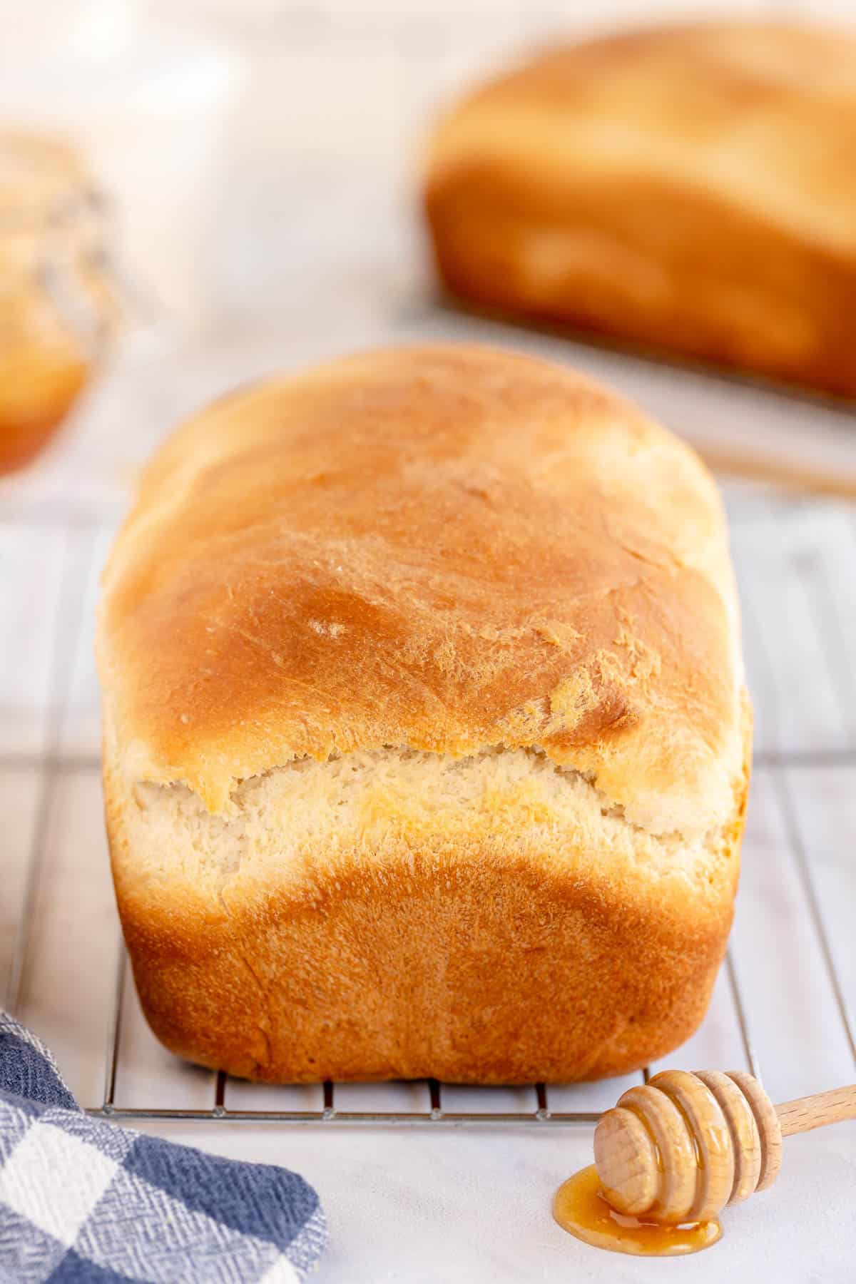 A freshly baked loaf of sandwich bread cooling on a wire rack is accompanied by a honey dipper with honey and a checked cloth in the foreground. Another loaf can be seen in the background.