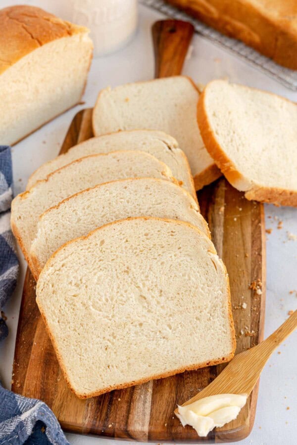 A wooden cutting board with four slices of sandwich bread, a loaf in the background, and a small wooden knife spread with butter nearby.