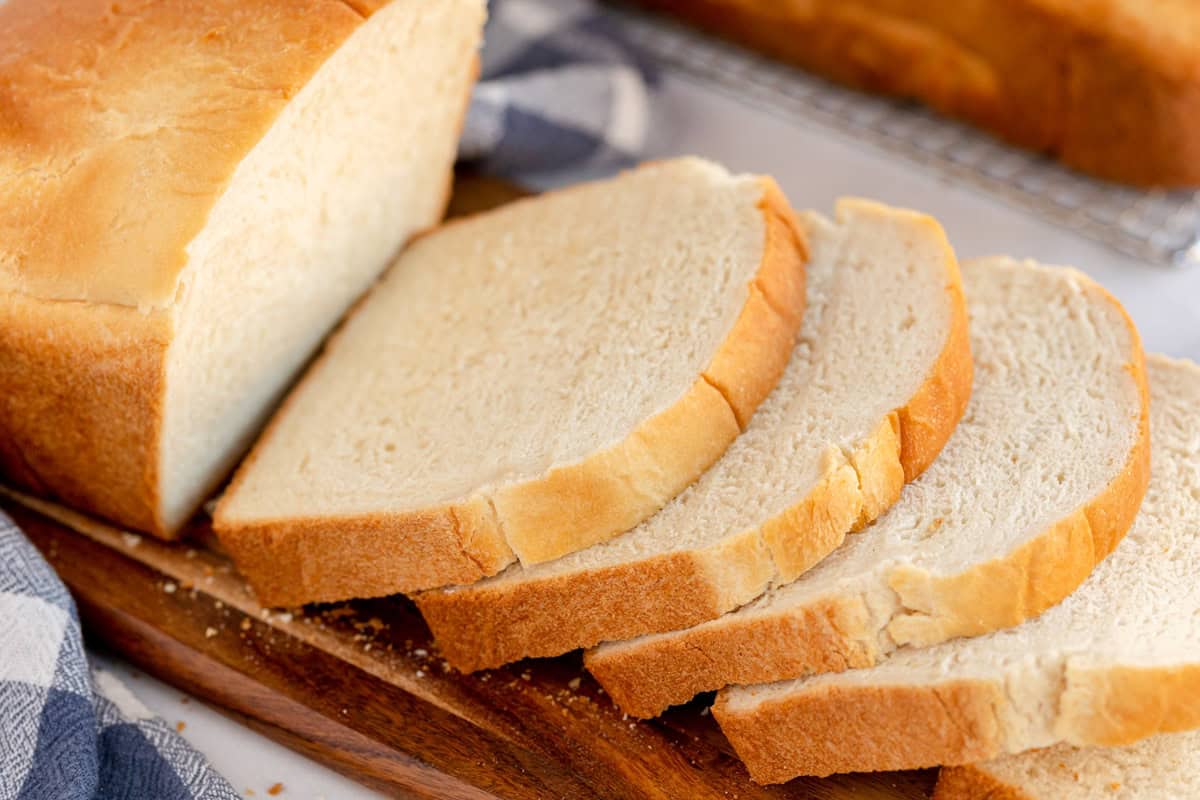 A loaf of sandwich bread partially sliced on a wooden cutting board, with slices fanned out and a striped cloth in the background.
