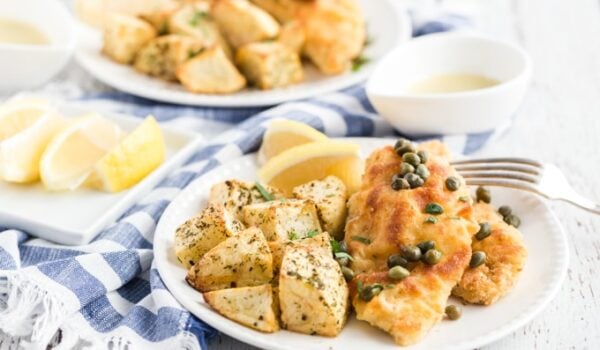 chicken piccata on a plate with roasted potatoes, and cut up lemon with a blue and white napkin
