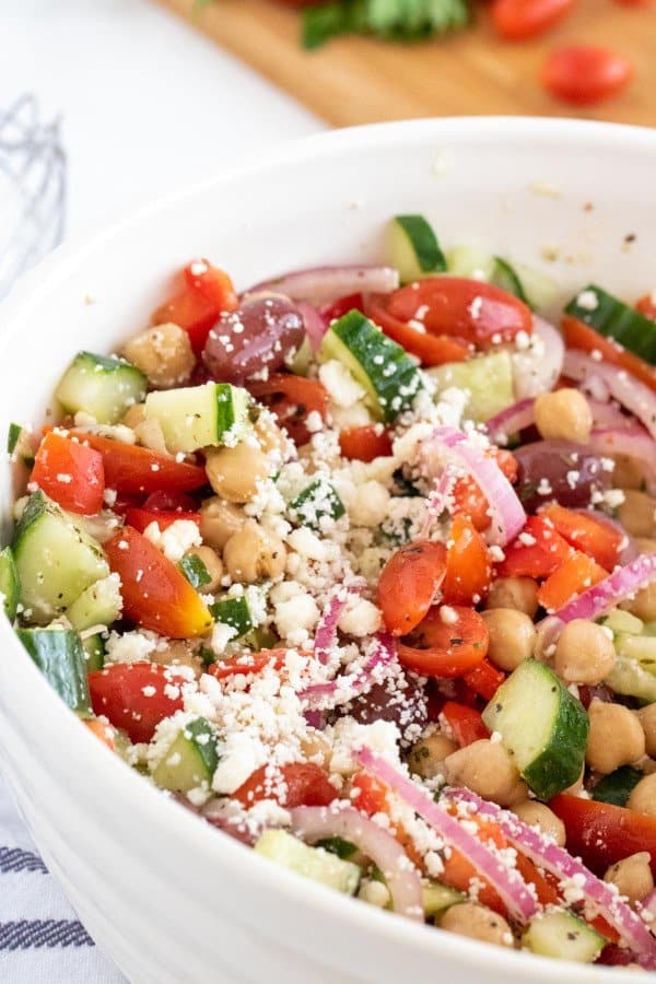 greek chick pea salad in a white bowl with cucumbers and tomatoes on a chopping board.