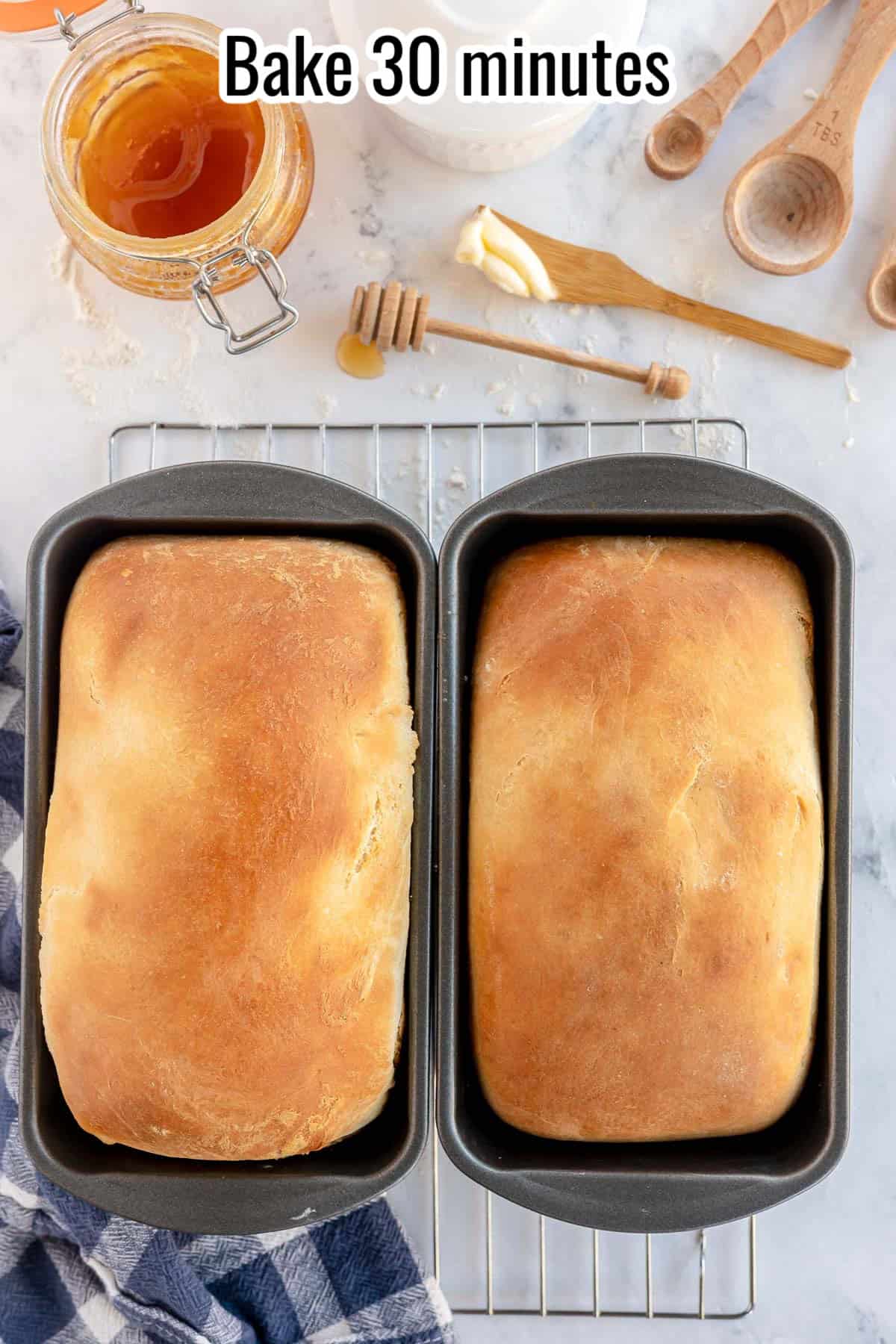 Two loaves of freshly baked bread in pans on a cooling rack. Wooden kitchen tools, a jar of honey, and a blue cloth are beside them. The text "Bake 30 minutes" is displayed at the top.