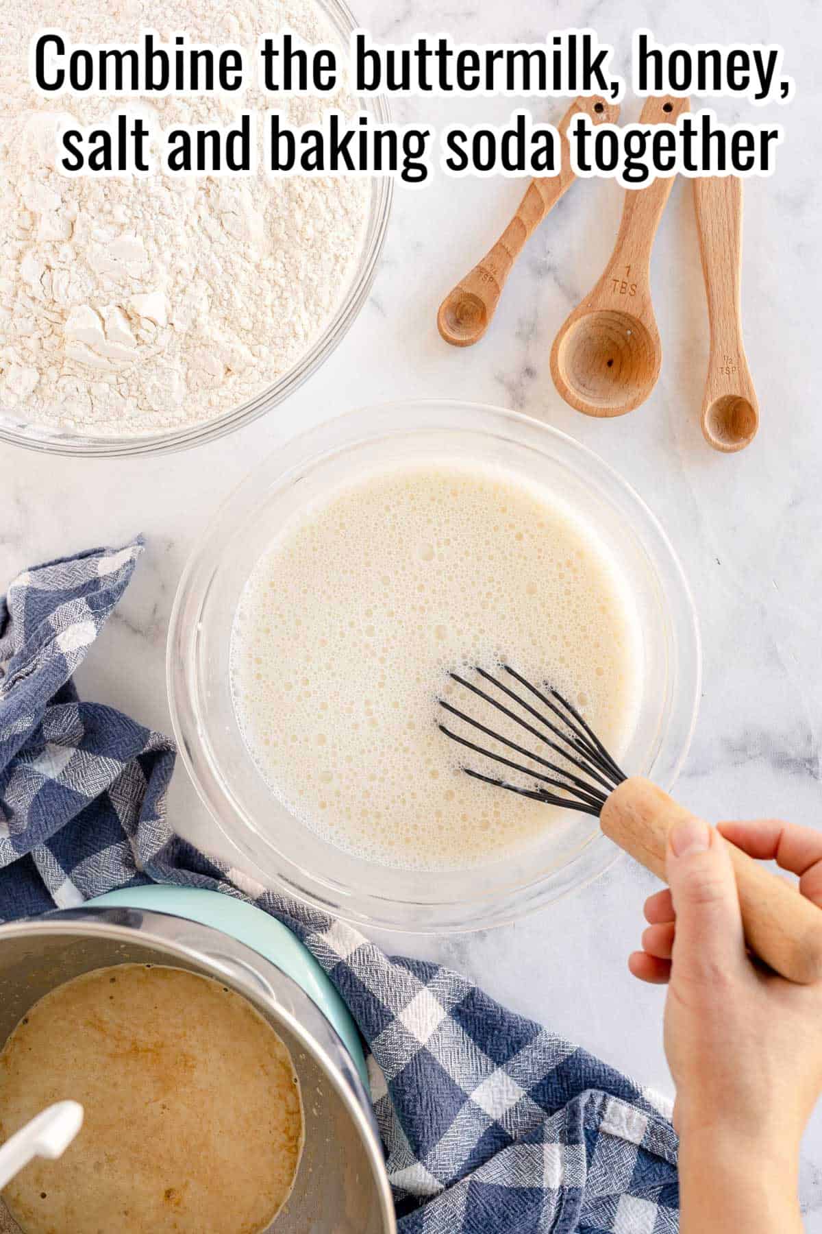 A bowl of mixed buttermilk, honey, salt, and baking soda is being whisked by hand to make sandwich bread. Flour and measuring spoons are on the side.