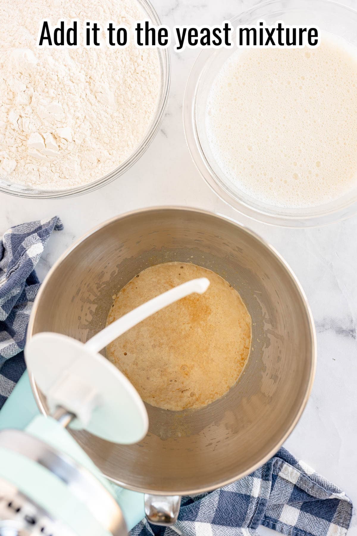 Overhead view of an ingredients setup for baking sandwich bread, with flour in a bowl, yeast mixture in another, and a stand mixer bowl containing a mixture. Text reads "Add it to the yeast mixture".