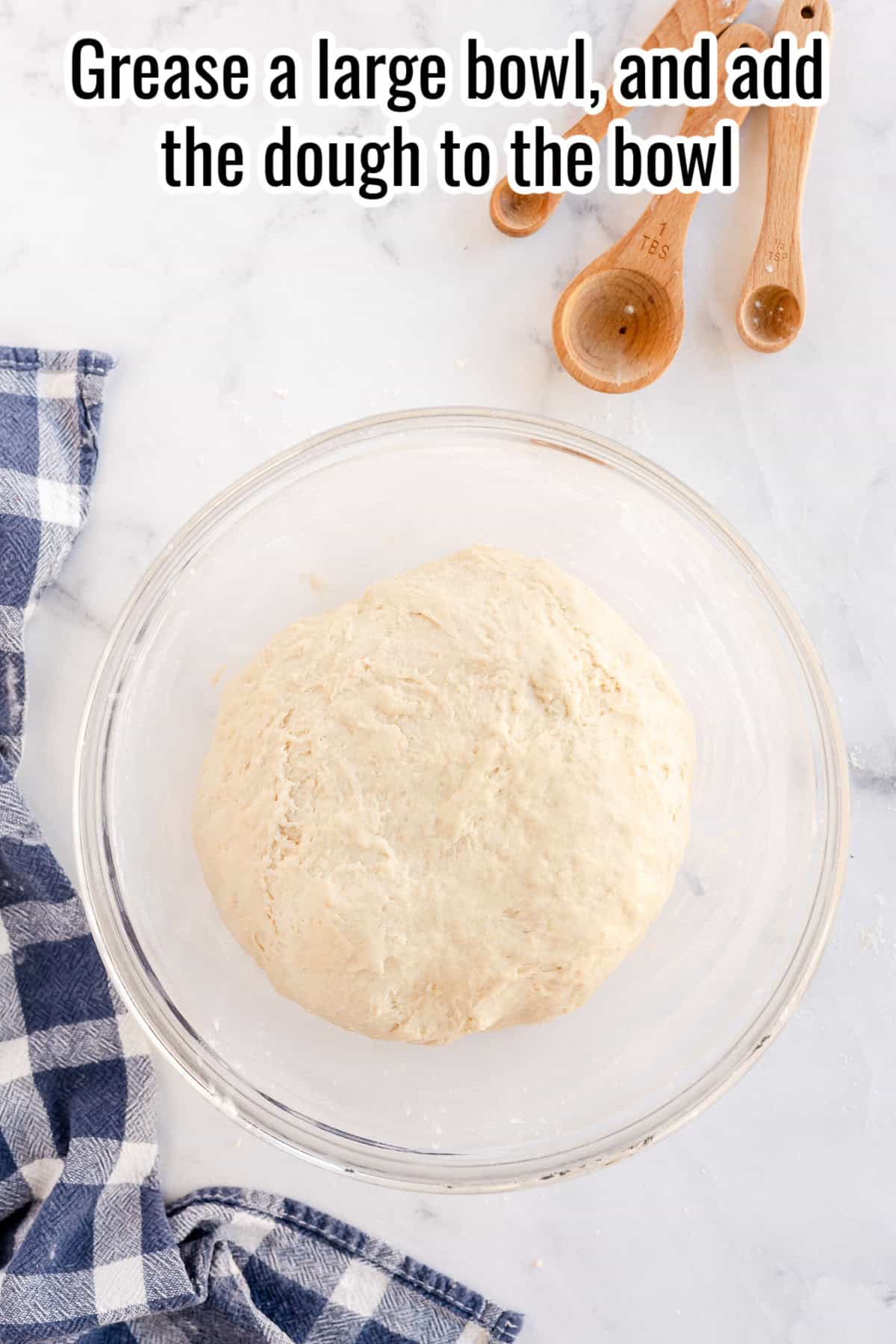 A ball of dough rests in a large glass bowl on a marble surface, with wooden measuring spoons and a checked cloth nearby. Text at the top reads, "Grease a large bowl, and add the dough for sandwich bread to the bowl".