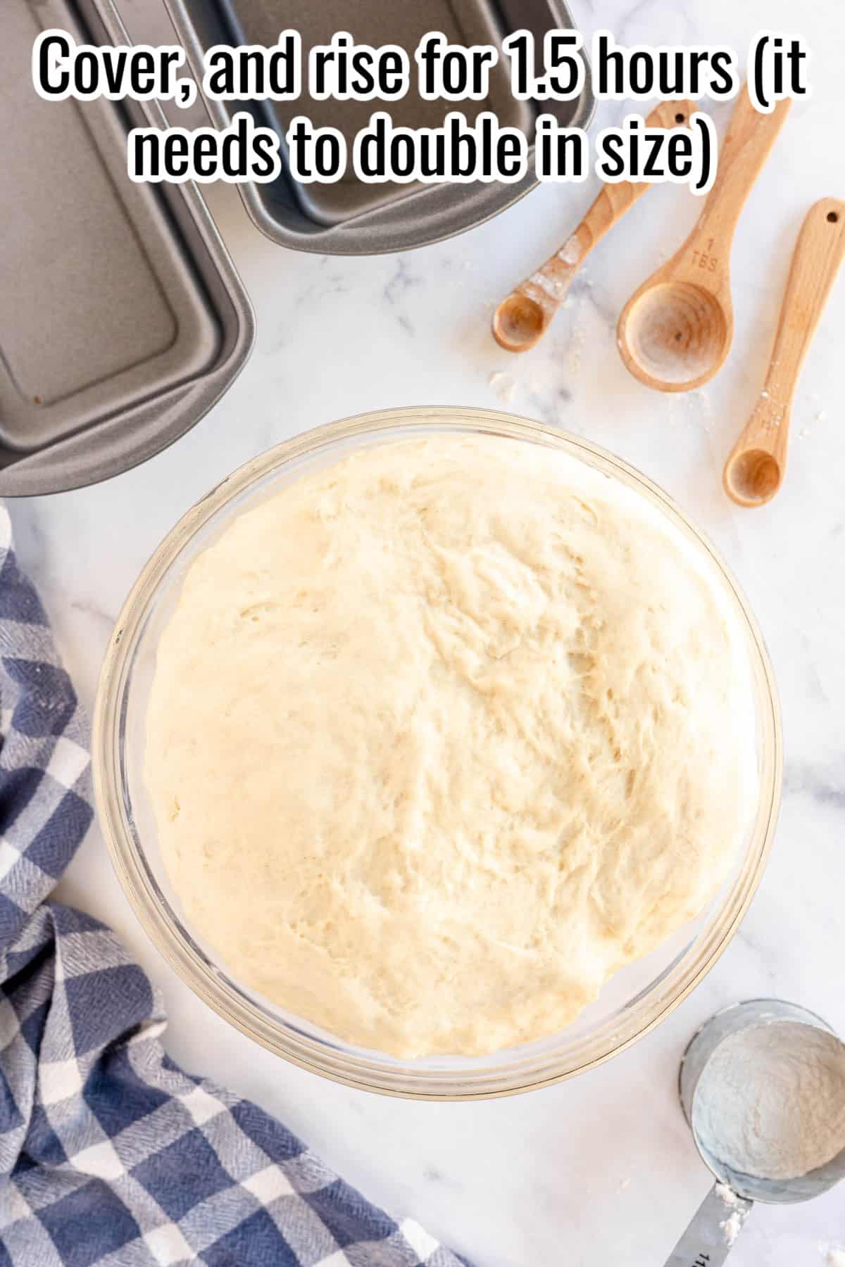 A bowl of bread dough, meant for sandwich bread, is placed on a countertop beside measuring spoons, a loaf pan, a kitchen towel, and some flour. Text on the image instructs to cover and let rise for 1.5 hours until it doubles in size".