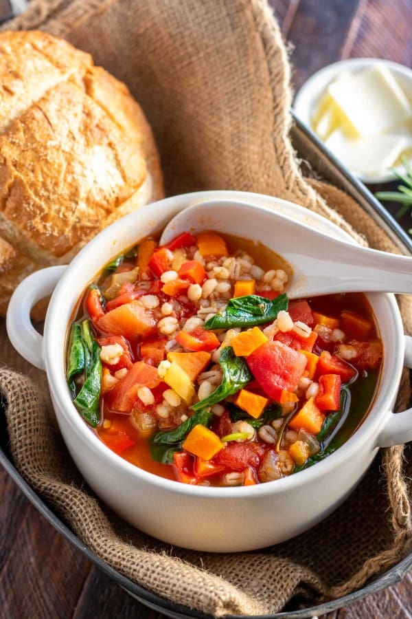 vegetable barley soup in a white bowl with a bread roll.
